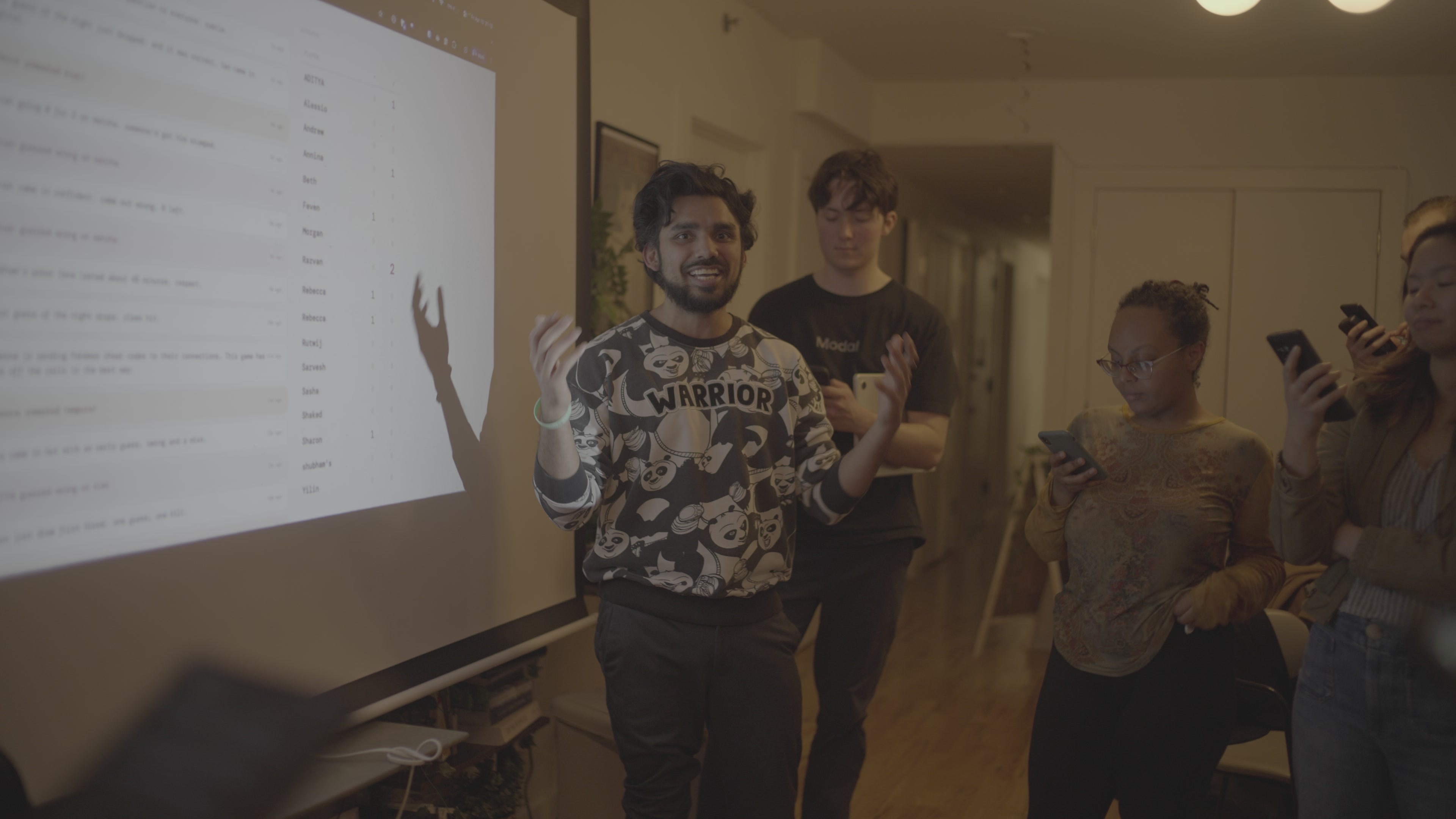 The room at the party, out of focus: the host beside a projected leaderboard, guests on their phones